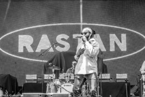 Male singer wearing a bandana and sunglasses performing on stage with a microphone; drummer and Yamaha drum kit visible behind him.