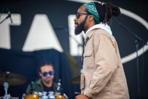 Side profile of a man in a teal bandana and sunglasses on stage, beige jacket, with a drummer blurred in the background.