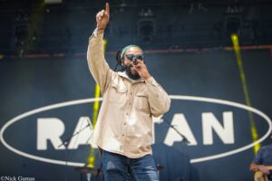Male singer on stage raises his right index finger while singing into a microphone, wearing a beige jacket and sunglasses against a dark backdrop.