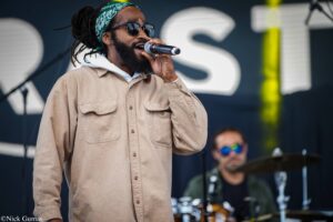 Male singer with dreadlocks and a green bandana, wearing sunglasses and a tan shirt, holding a microphone on stage with a drummer in the background.