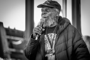 Older man with a gray beard speaks into a handheld microphone at an outdoor event, wearing a cap and puffer jacket .
