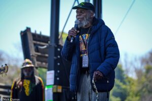 Older Black man with gray beard wearing a navy jacket and green cap speaks into a microphone on an outdoor stage.