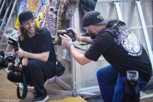Two videographers filming outdoors, one with a large camera rig and long hair, the other aiming a camera.