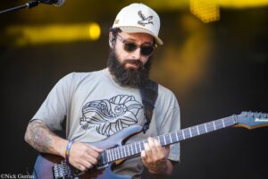 Bearded guitarist wearing sunglasses and a white cap, playing an Ibanez electric guitar on stage.