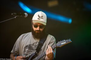 Bearded guitarist wearing a light cap with a bird logo and sunglasses, playing an electric guitar on stage.