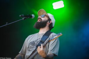 Male guitarist with a beard and baseball cap singing into a microphone on stage, green spotlight behind him