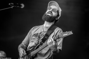 Male guitarist with beard, sunglasses, and cap, singing into a mic while playing an electric guitar on stage (black and white)