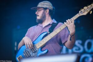 Bearded musician wearing a patterned cap and glasses plays a blue bass guitar on stage.