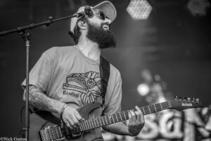Bearded guitarist wearing a hat and sunglasses, playing a double-neck Ibanez guitar on stage.