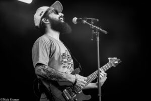 Bearded guitarist wearing a cap and sunglasses, singing into a microphone while playing an electric guitar on stage (black and white).