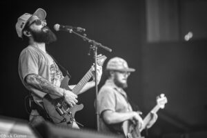 Bearded guitarist in cap and sunglasses sings into a microphone while playing a solid-body electric guitar on stage (black and white).]