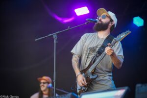 Male guitarist with a beard and cap playing an electric guitar on a lit stage, purple and blue lights in the background.
