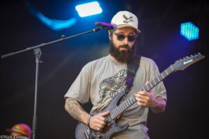 Bearded guitarist in sunglasses and a beige cap playing an Ibanez electric guitar on stage with a microphone stand nearby.