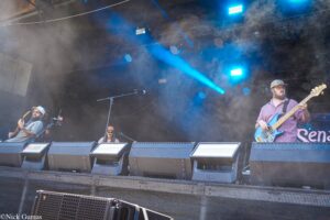 Band performing on an outdoor stage with blue lights and fog; a guitarist on the left and a bassist on the right.