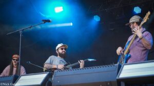 Band on stage performing with guitars under blue lights; guitarist wearing cap and beard, bassist in pink shirt, and other guitarist in sunglasses and hat.