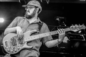 Black-and-white photo of a bearded musician playing a bass guitar on stage, wearing a patterned cap and glasses.