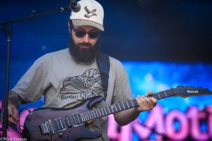 Bearded guitarist wearing a cap and sunglasses playing a brown Ibanez electric guitar on stage during a live performance.