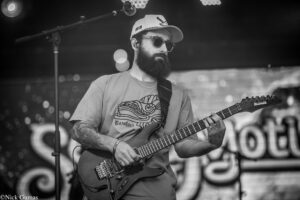 Bearded guitarist in sunglasses and cap playing an Ibanez electric guitar on stage.