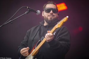 Male guitarist in sunglasses and a black hoodie performs on stage, playing an electric guitar near a microphone.