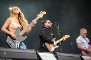 Female bassist with blond hair playing a gray bass guitar on stage, with a singer and another guitarist in the background.