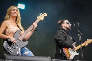 Female bassist with blonde hair playing a gray bass on stage, wearing a beige top; a male singer in sunglasses sings into a mic in the background.