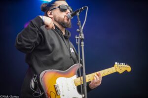 Guitarist wearing sunglasses and a black hoodie plays a sunburst Fender Stratocaster on stage, singing into a microphone.