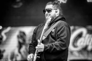Guitarist with beard and sunglasses playing an electric guitar on stage, smiling in a black-and-white photo.