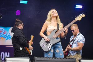Smiling female bassist playing a gray bass guitar at center stage, flanked by two guitarists during a live outdoor performance.