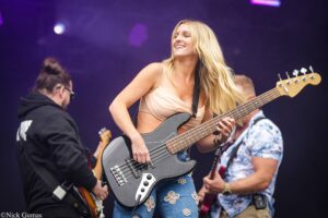 Female bassist smiling while playing a gray electric bass on stage with purple lights.