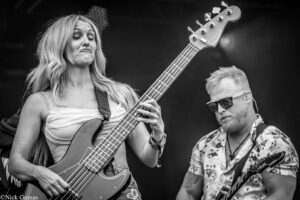 Black-and-white photo of a female bass guitarist with long blonde hair playing a bass on stage, a male guitarist beside her wearing sunglasses on the right.