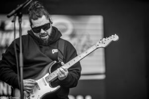 Male guitarist wearing a hoodie and sunglasses, playing an electric guitar on stage (black and white).