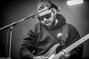 Black-and-white photo of a bearded guitarist wearing sunglasses and a hoodie, playing an electric guitar into a microphone during a live performance.