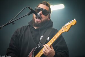 Male guitarist and vocalist wearing sunglasses and a black hoodie, singing into a microphone on stage with an electric guitar visible in the foreground.