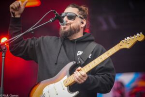 Male guitarist wearing sunglasses and a black hoodie performs on stage, singing into a microphone while playing a sunburst Fender Stratocaster.