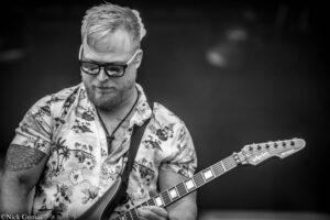 Black-and-white photo of a bearded man wearing sunglasses and a tropical shirt, playing an electric guitar.