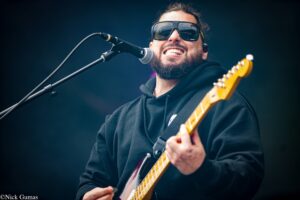 Bearded guitarist wearing oversized sunglasses and a black hoodie, smiling into a microphone while playing a cream-colored electric guitar on stage.