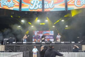 Outdoor concert stage at a festival with a live band performing under bright yellow stage lights and colorful banners above the crowd barrier.