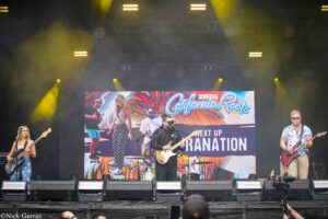 Band performing on an outdoor stage with a colorful 'California Roots' banner in the background.