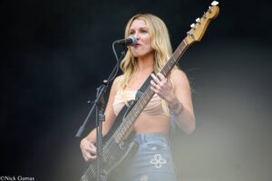 Female bassist singing into a microphone on stage while playing a bass guitar outdoors.