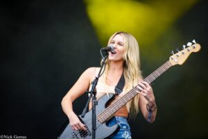 Female bassist singing into a microphone on stage while playing a gray bass guitar with a strap, in casual clothes and jeans.
