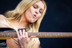 Close-up of a blonde woman singing while playing an acoustic guitar, lips puckered near the strings and fretboard visible.