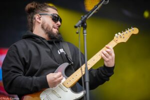 Musician wearing sunglasses and a black hoodie plays a Fender Stratocaster on stage with a microphone stand nearby.