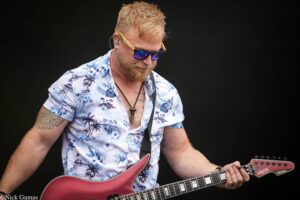 Male guitarist performing on stage, wearing a blue floral shirt and sunglasses, playing a double-neck red guitar against a dark background