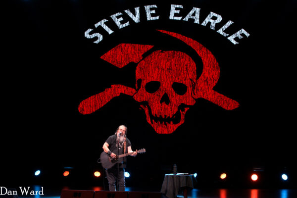 Guitarist performs on stage under blue and orange lights with a large red skull banner behind him.