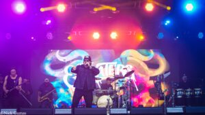 Lead singer in a denim jacket and cap performing on a brightly lit stage with a colorful psychedelic backdrop and band members playing instruments behind him.