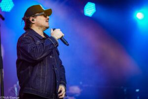 Male singer wearing a denim jacket, cap, and sunglasses, holding a microphone on stage under blue lights.