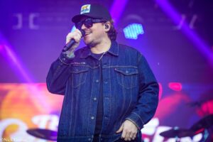 Male singer wearing a denim jacket and cap with a logo, holding a microphone on stage with purple lighting in the background.] ,**Oops**