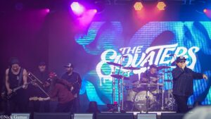 Live concert scene: musicians on stage with guitarist, bassist, drummer; purple and blue lights and a large leafy backdrop behind them.