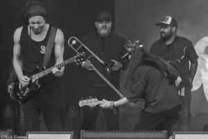 Guitarist in a black sleeveless shirt plays an electric guitar on stage as two other musicians with brass instruments stand nearby in a live performance.