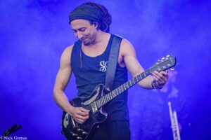Musician with dreadlocks and headband playing a dark electric guitar on a blue-lit stage.
